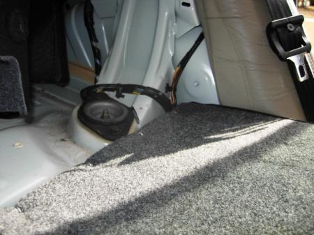 Left side shock mount on sedan located behind rear seat under cover.